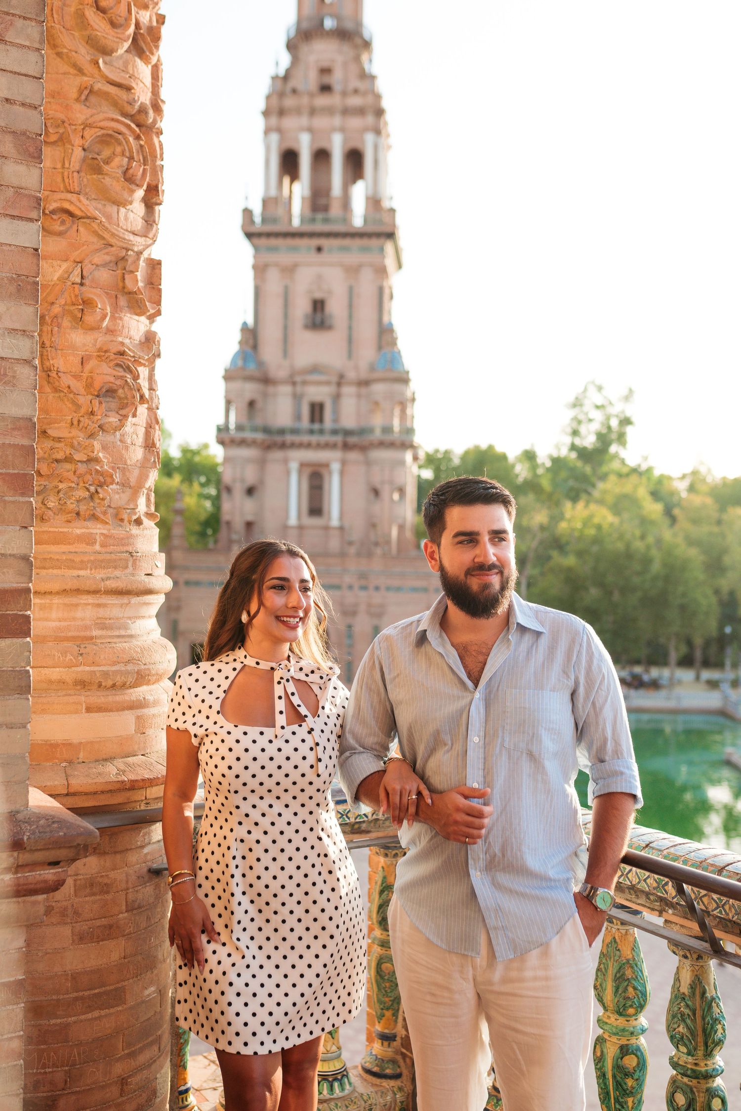 Couple smiling at Plaza de España in Seville, Spain, with ornate tower and canal in background.