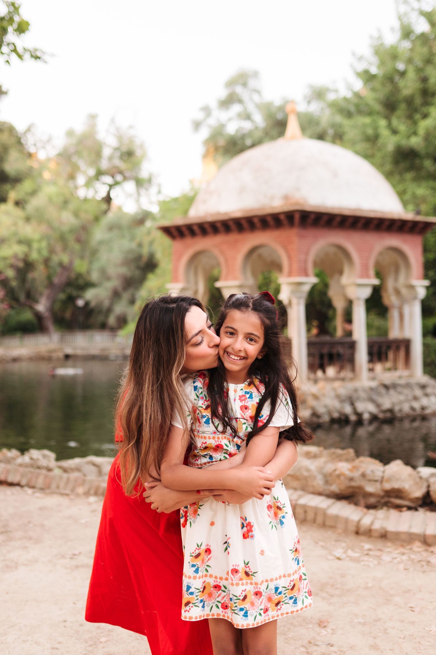A woman in red dress hugging and kissing a smiling girl in floral dress near a decorative pavilion by a pond.