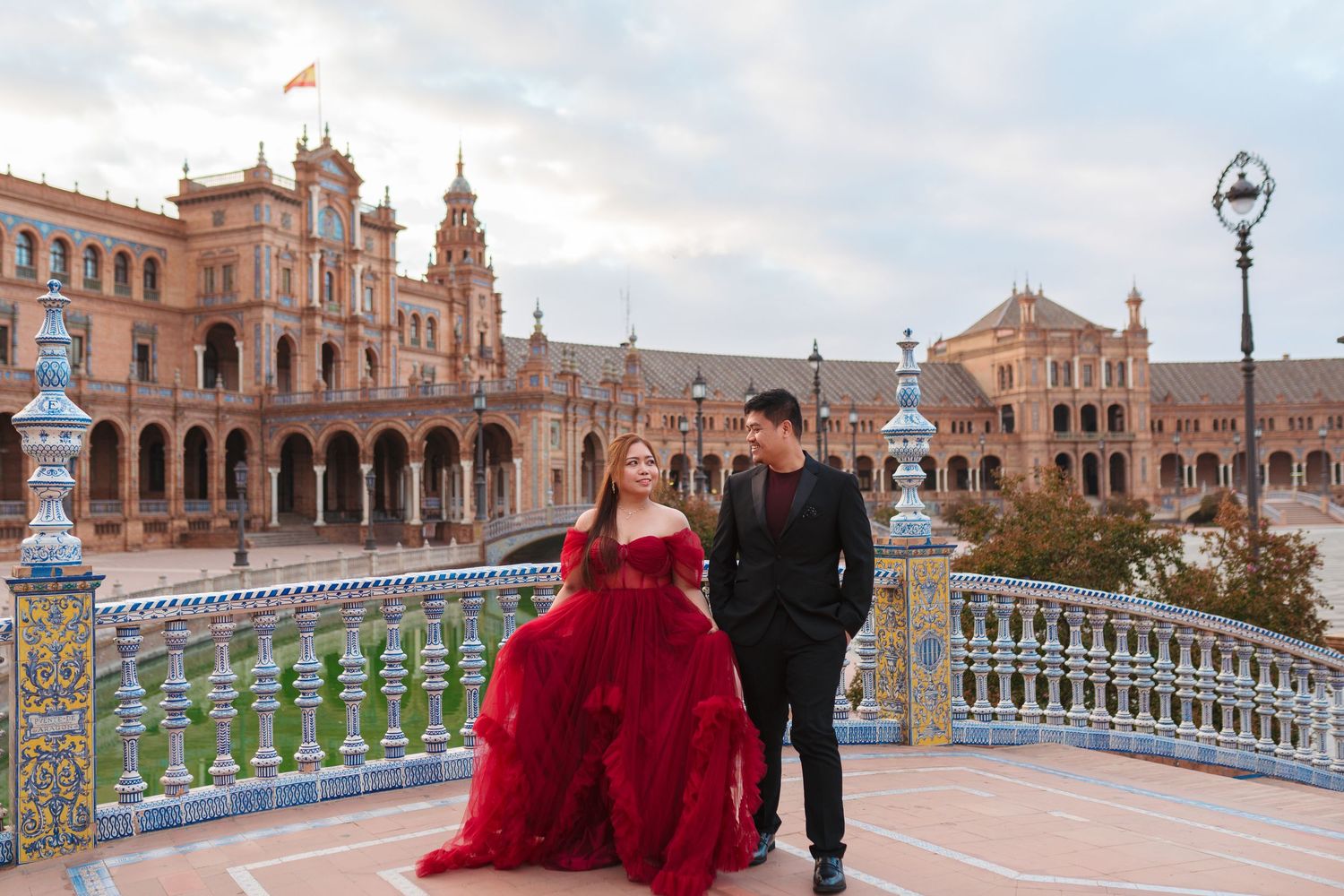 Couple in elegant attire poses at Plaza de España, Seville, woman in red gown, man in black suit.