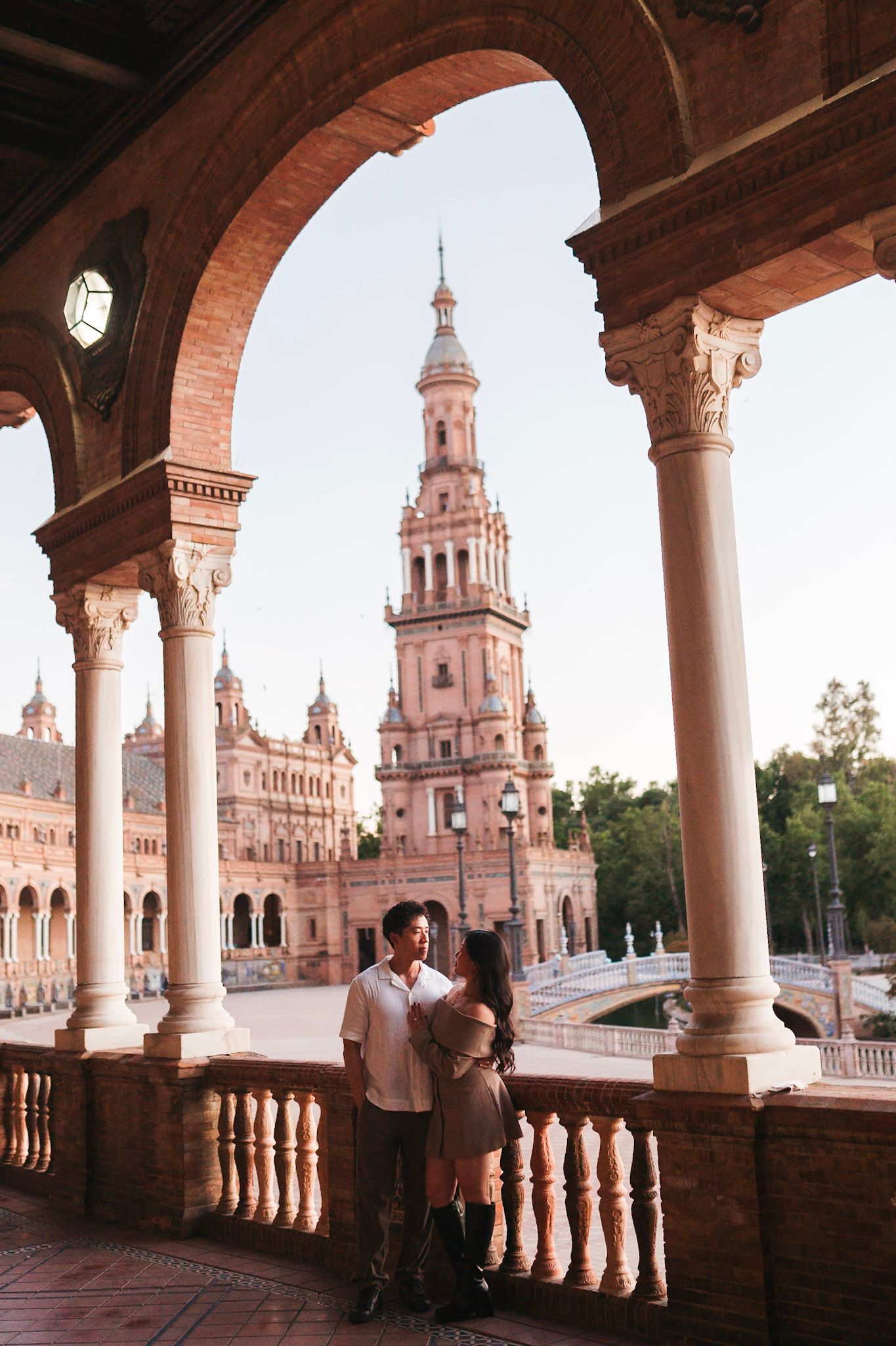 Couple embracing under ornate arches at Plaza de Espana with tower backdrop in Seville.