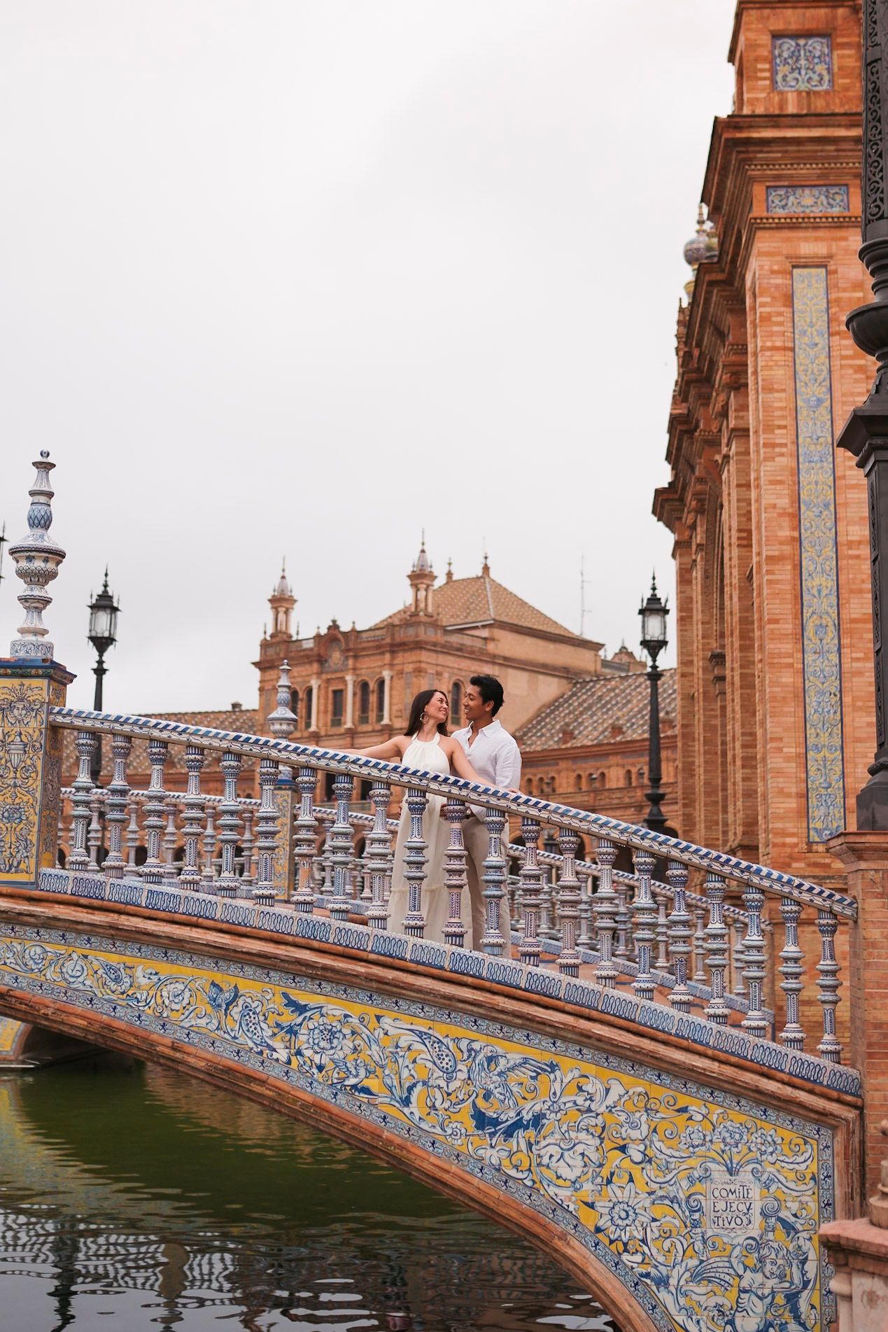 Couple embracing on colorful mosaic-tiled bridge over canal at Plaza de España, Seville, Spain.