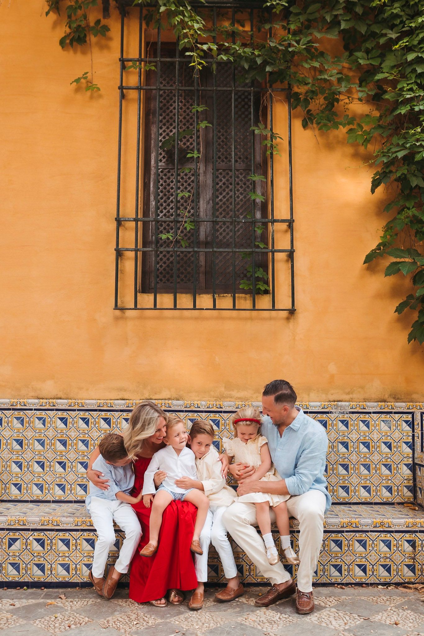 Family of six sitting together on a decorative tiled bench against a yellow wall in Seville.
