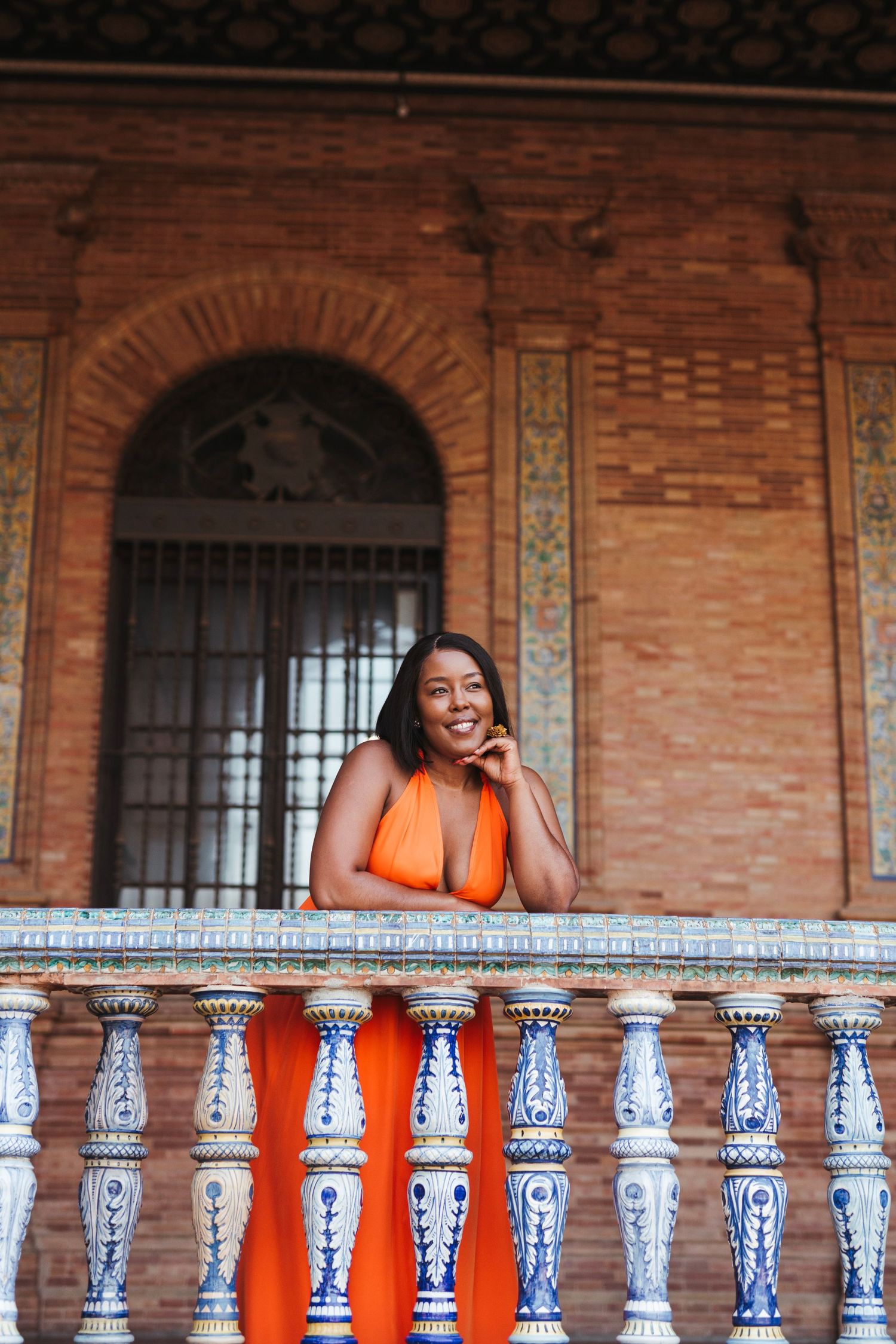 Smiling woman in orange dress leaning on blue and white ceramic balustrade at Plaza de España, Seville.