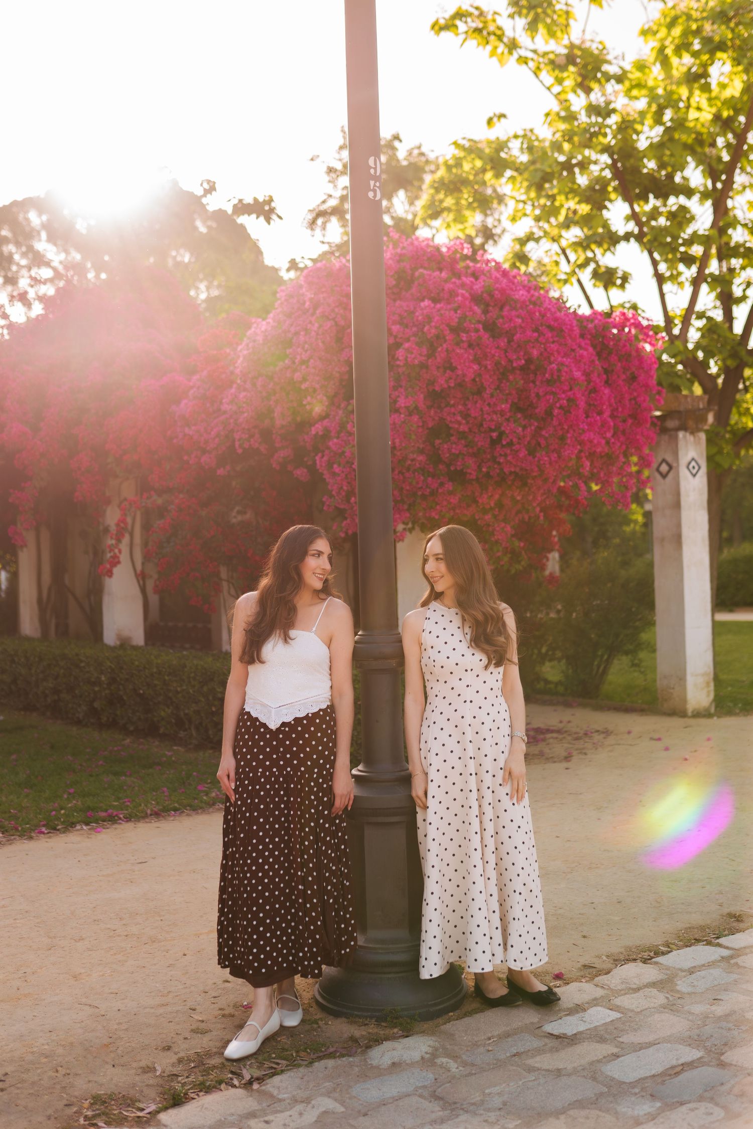 Two young women in polka dot dresses stand by a lamp post with pink flowering trees in golden sunlight.