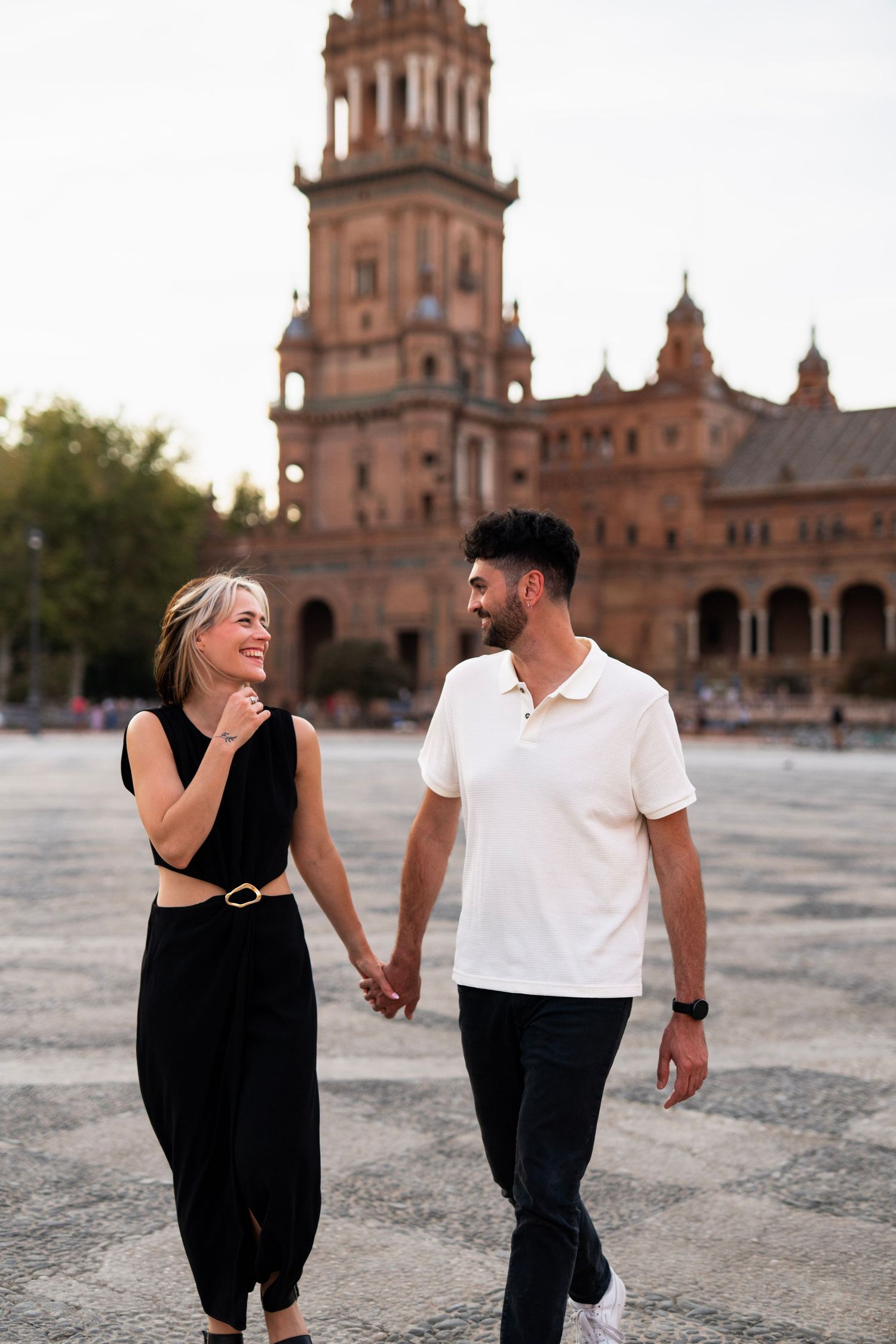 Smiling couple holding hands walking across Plaza de Espana with tower in background.