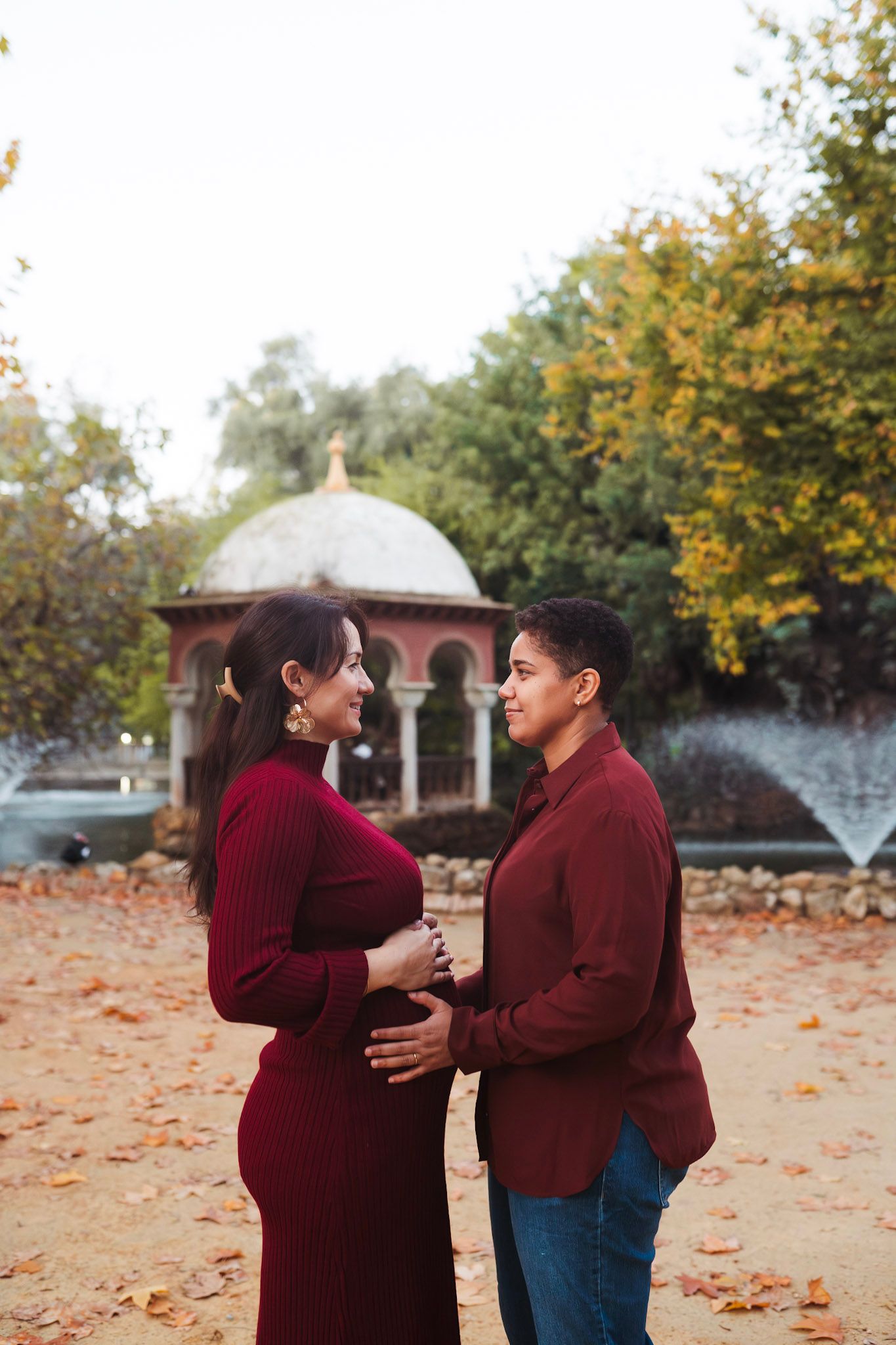 Pregnant woman in burgundy dress and partner share tender moment by fountain in autumn park.