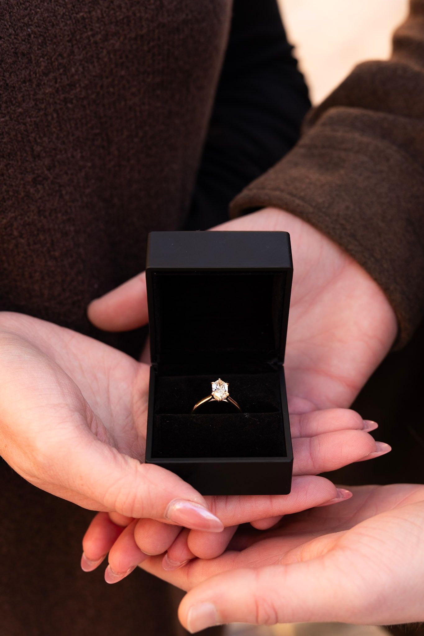 Two hands holding open black velvet box with solitaire diamond gold engagement ring together.