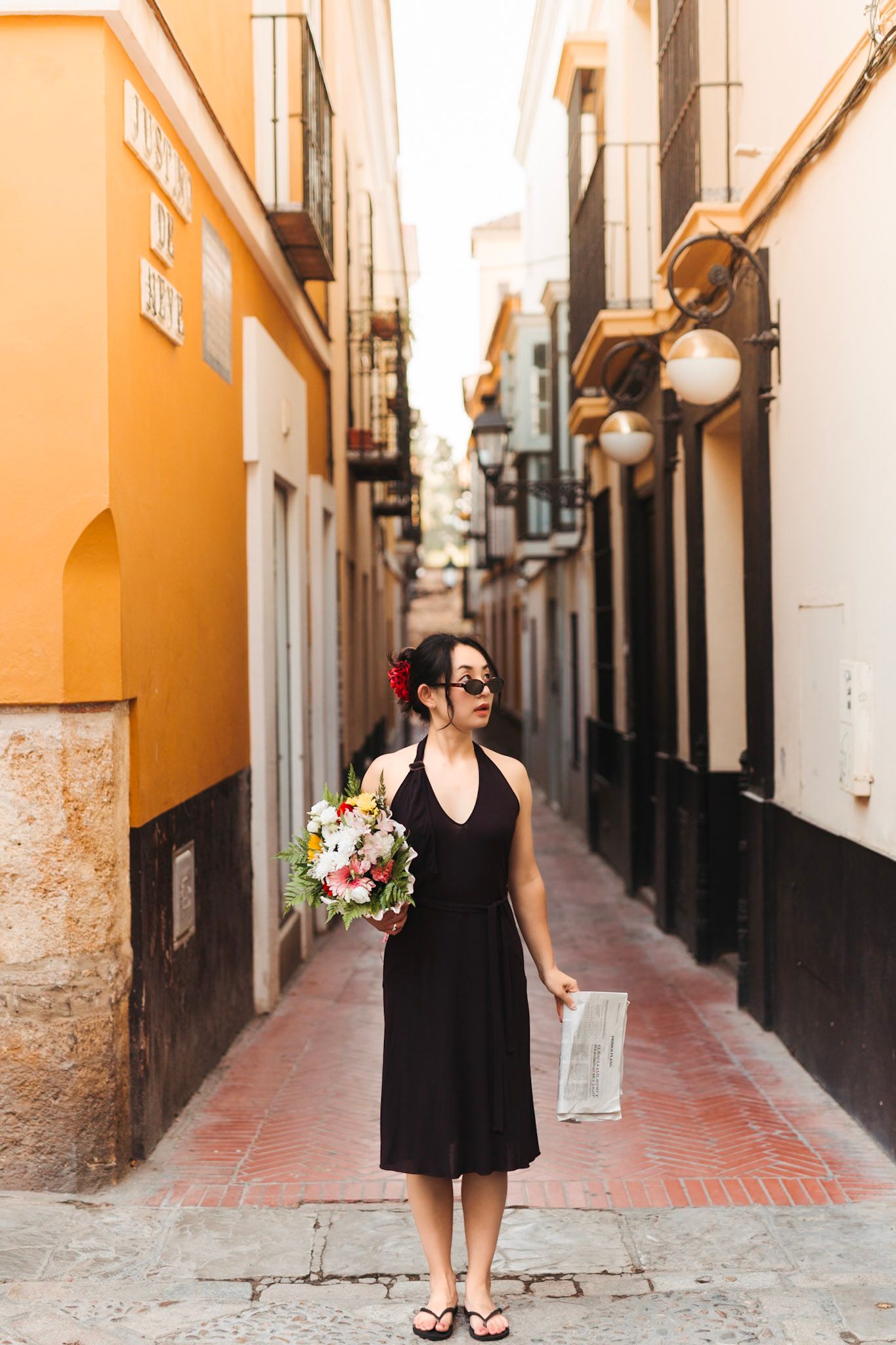 Asian woman in black halter dress holding bouquet standing in narrow Spanish street at golden hour.
