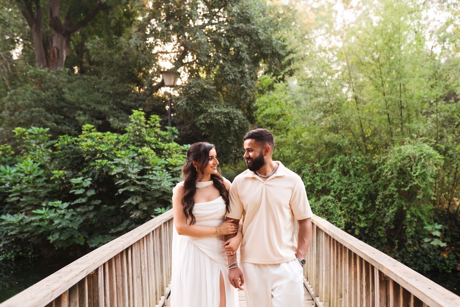 Couple in white outfits standing on wooden bridge surrounded by lush green trees, sharing a romantic moment.