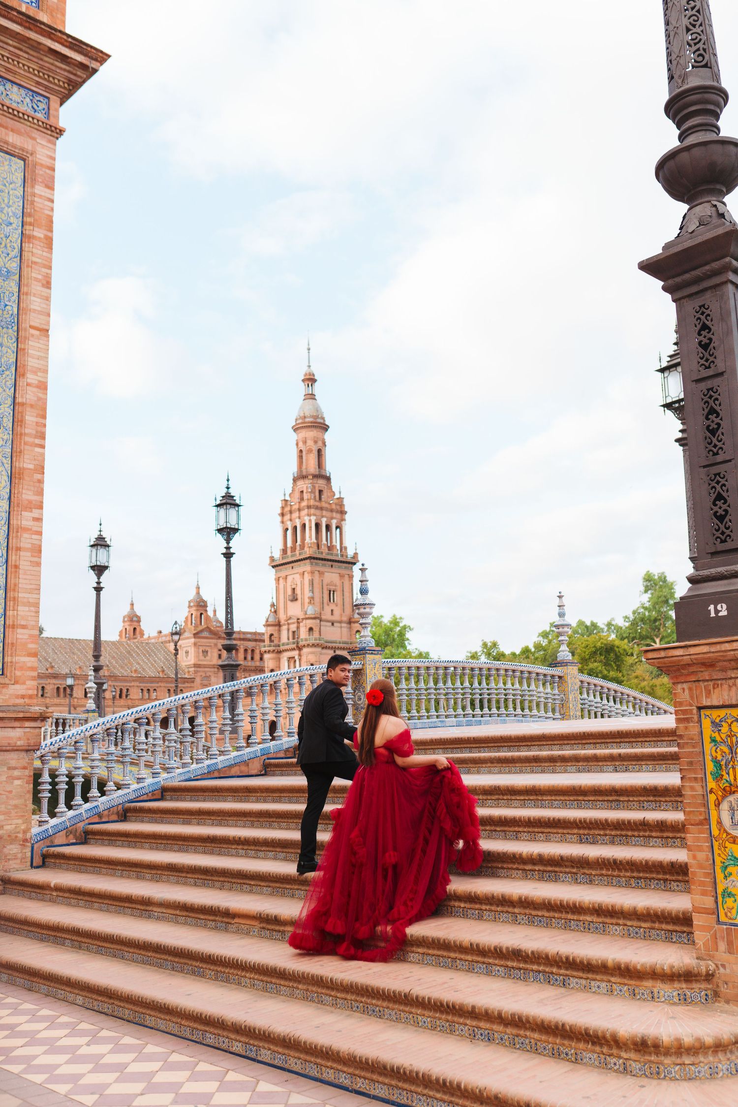 Couple in elegant attire on ornate steps at Plaza de España, Seville, woman in red gown, historic tower backdrop.