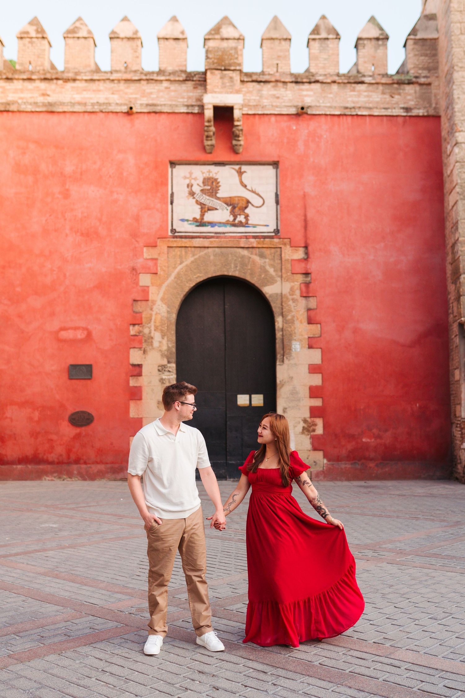 Couple holding hands in front of a red Moorish wall with lion emblem, woman in flowing red dress, Seville Spain.