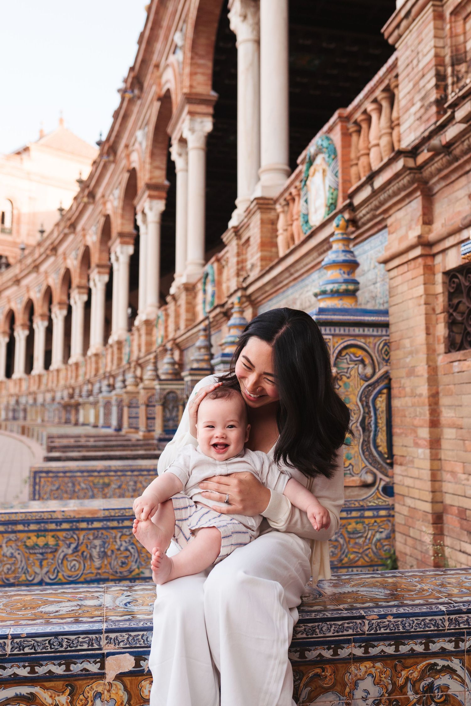 A mother smiles at her laughing baby on ornate tiled benches at Plaza de España, Seville, Spain.