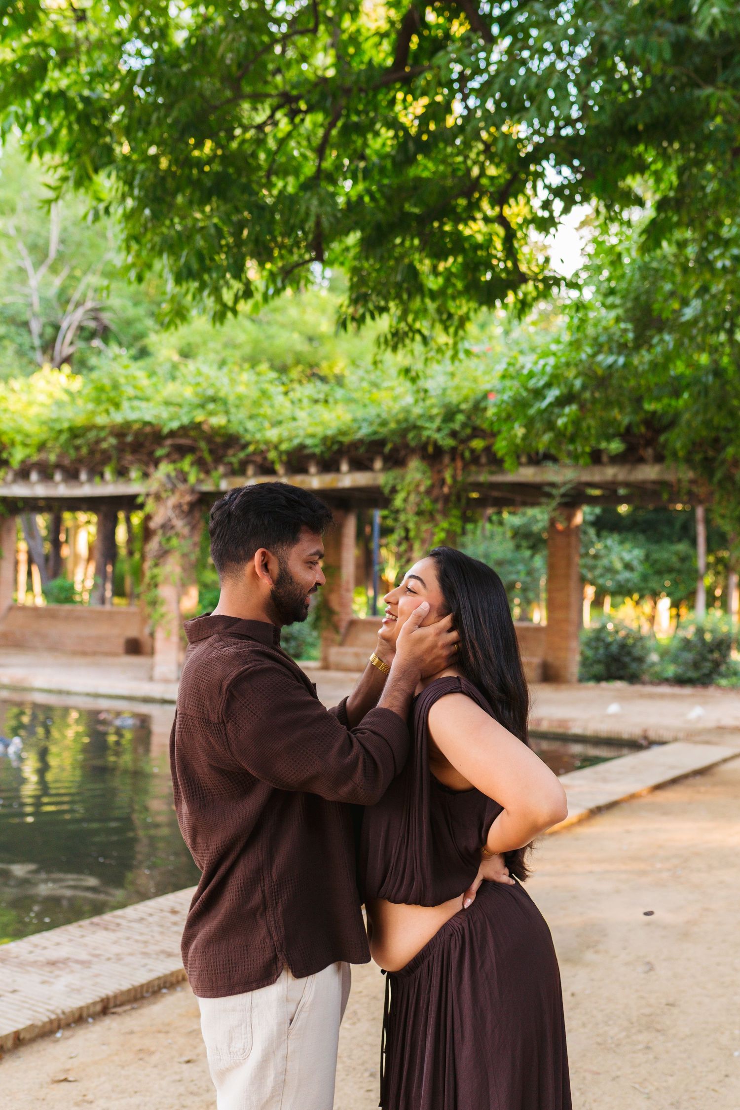 A couple shares a romantic moment in a lush green park, the man tenderly holding the woman's face as she smiles.