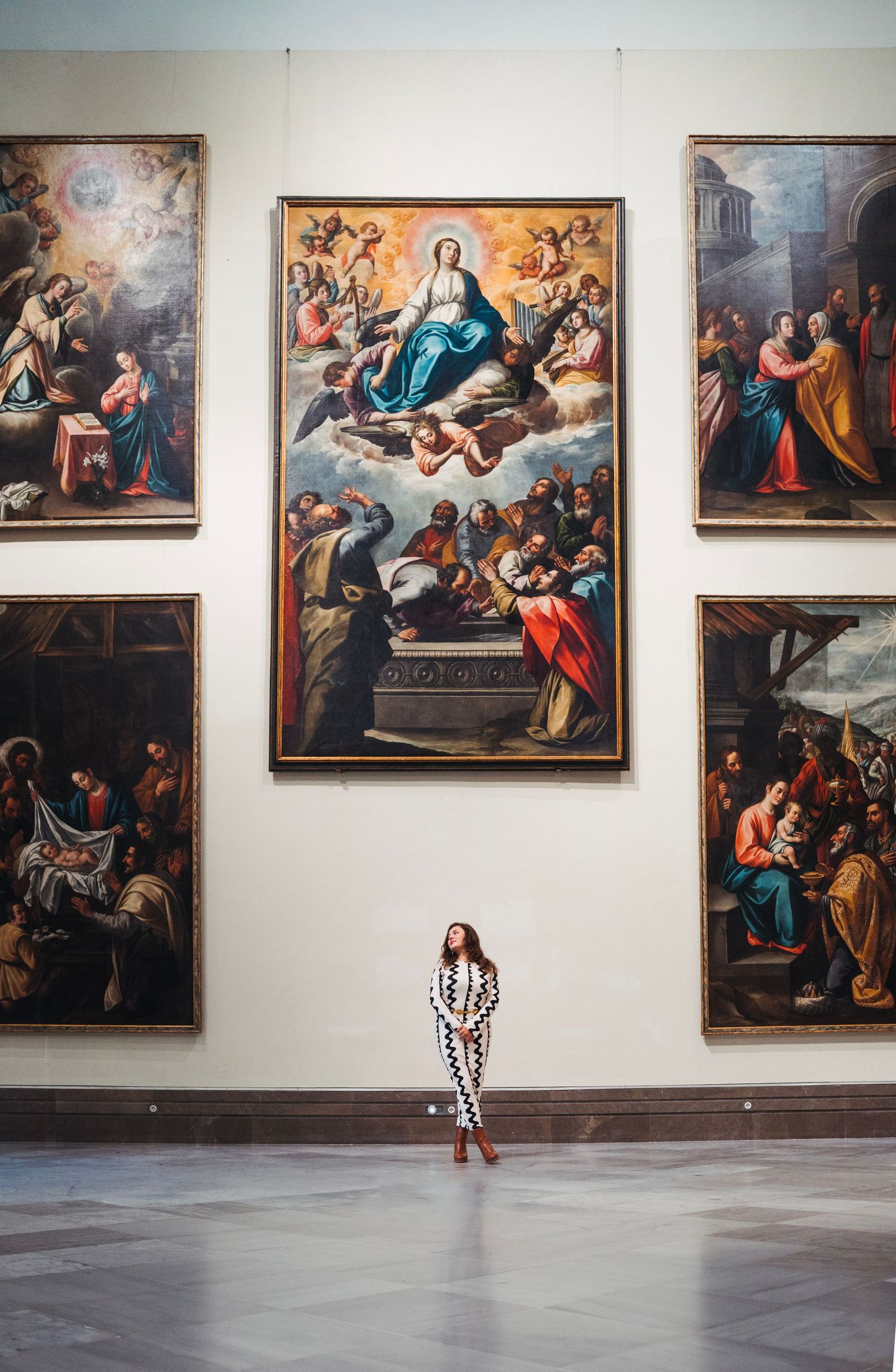 Woman in patterned outfit stands small beneath large classical religious paintings in a Seville museum.