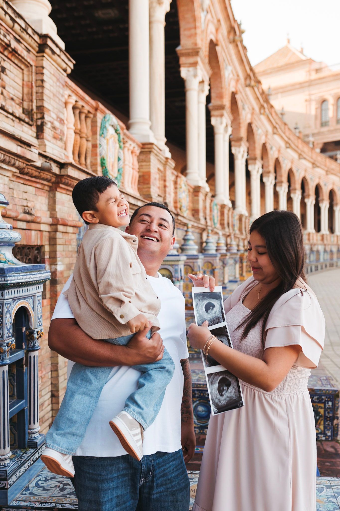A happy family at Plaza de Espana in Seville sharing pregnancy ultrasound photos outdoors.