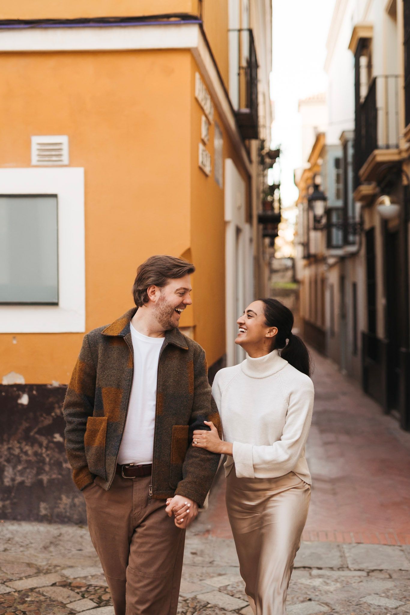 Happy couple walking hand in hand laughing through charming narrow street in Seville Spain.