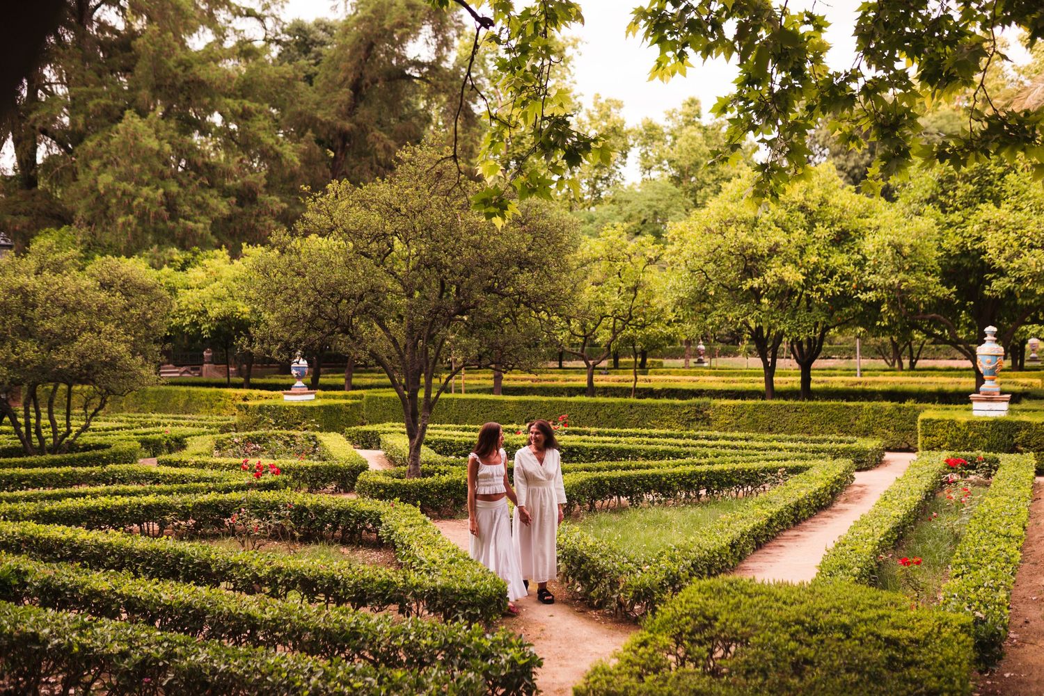 Two women in white dresses walk through an elegant formal garden maze with lush green hedgerows and trees.