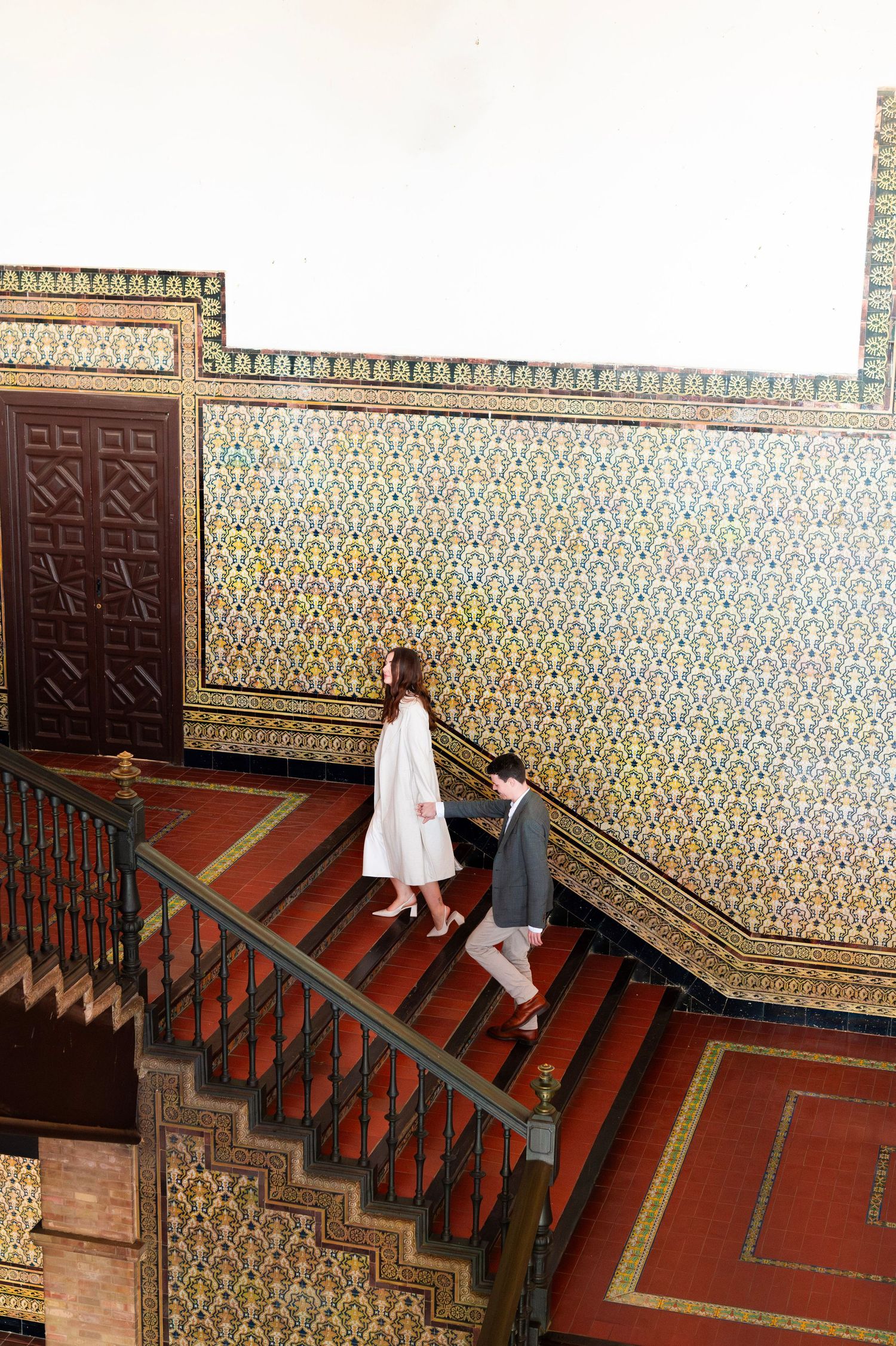 Couple walking down ornate mosaic-tiled staircase inside a historic Seville building, viewed from above.