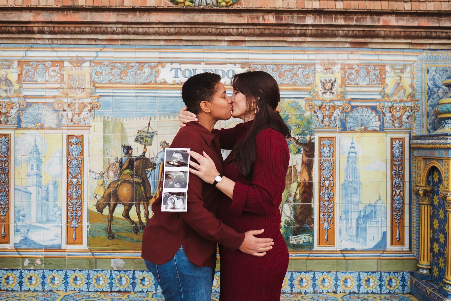 Expecting couple shares a kiss while holding ultrasound photos before vibrant Spanish tile murals.