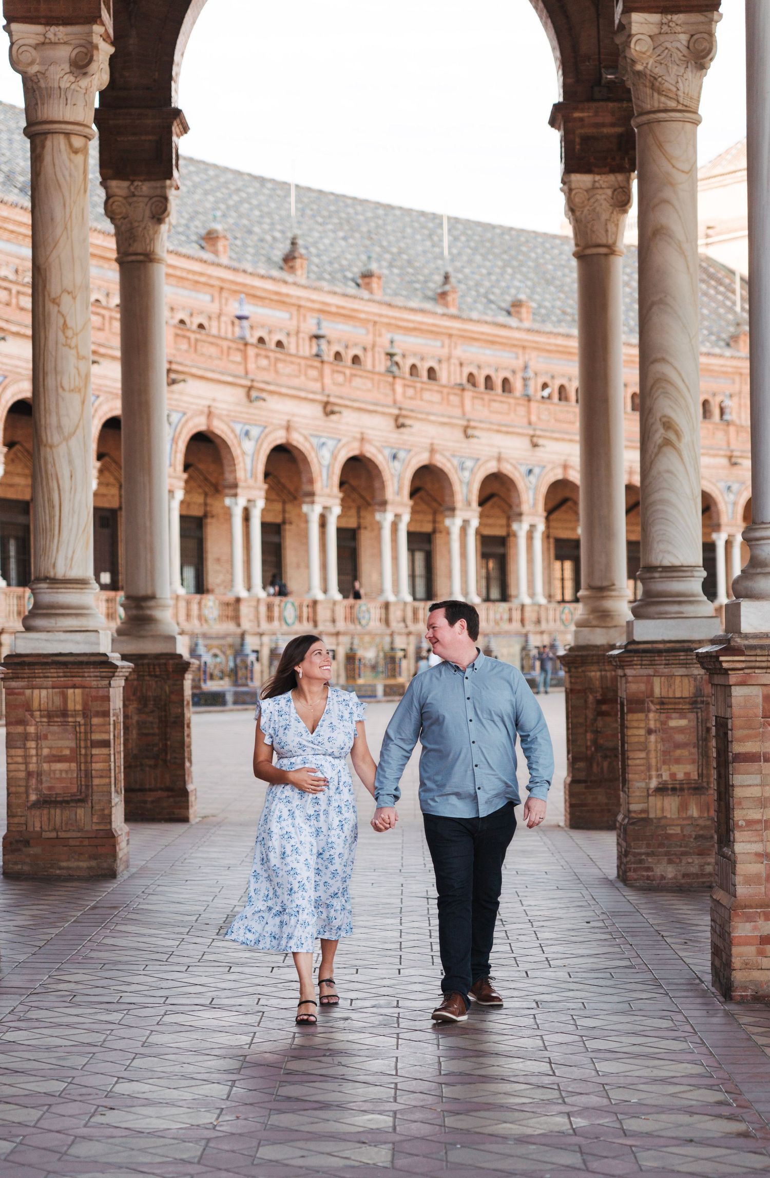 Pregnant couple walks hand in hand under arches at Plaza de España, Seville.