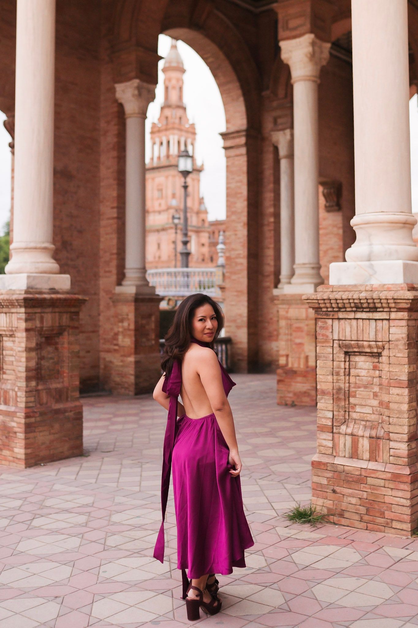 Woman in backless magenta dress walks through arched colonnade at Plaza de España, Seville, smiling over shoulder.