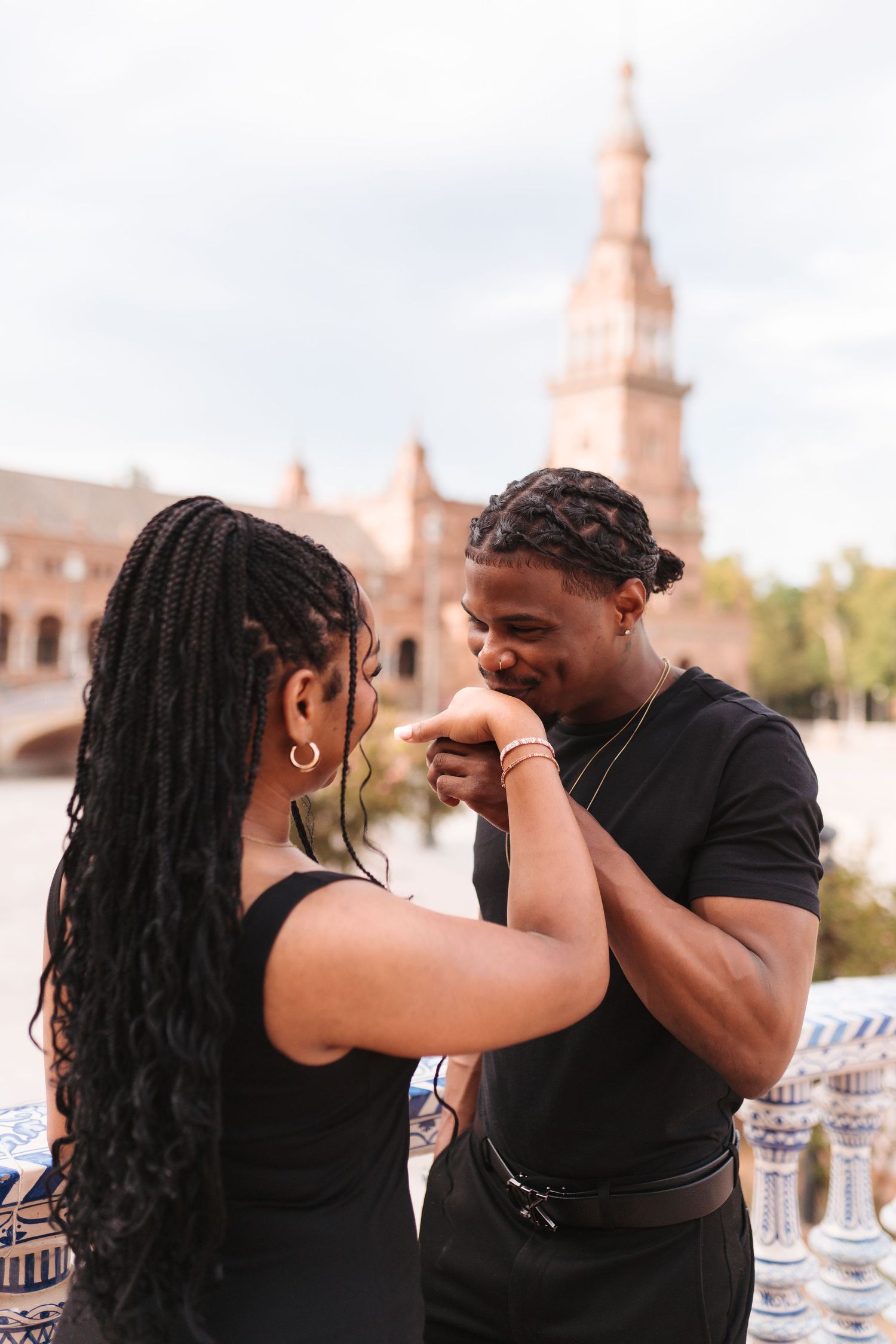 A romantic couple in black outfits share a tender moment at Plaza de España, Seville, as the man kisses the woman's hand.