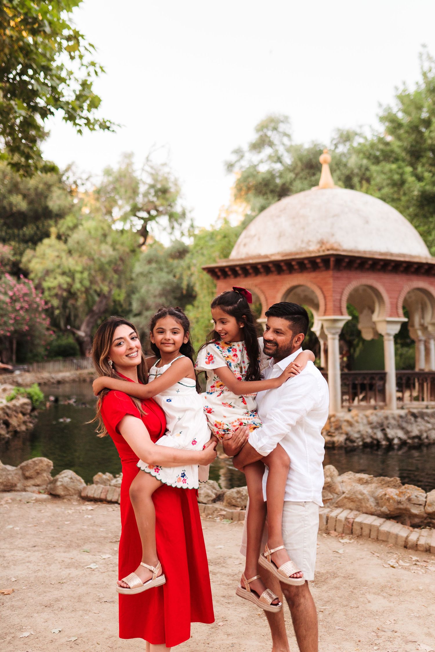 Happy family of four posing in a lush garden with a domed pavilion, parents in red and white holding two young daughters.