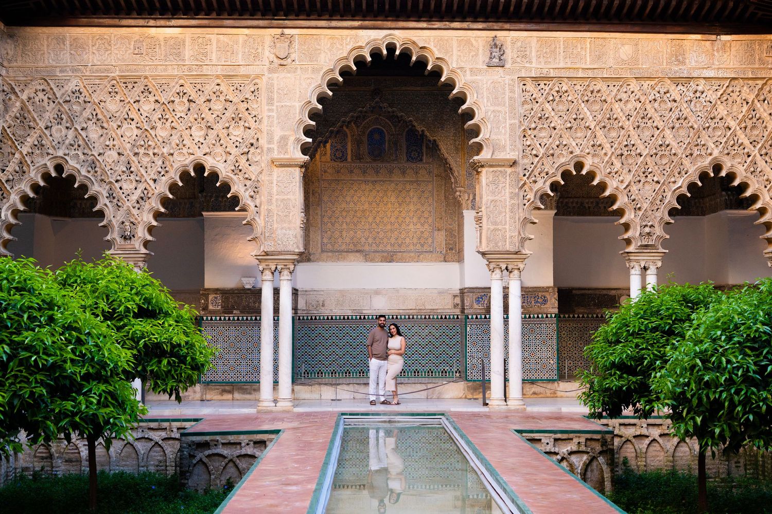 Couple standing by reflective pool in ornate Moorish courtyard with intricate carved arches in Seville.