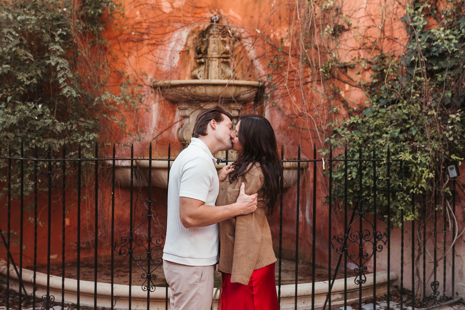 Couple sharing a kiss in front of a rustic fountain with ivy-covered orange walls.