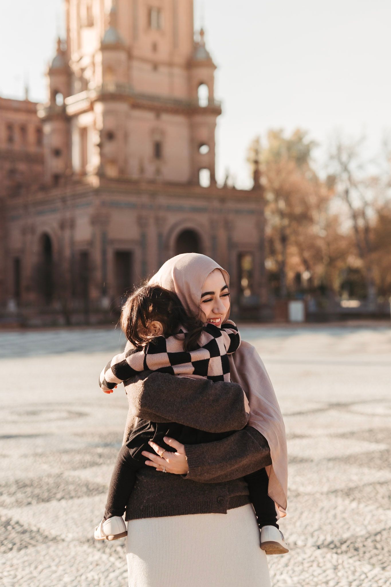 A woman wearing a hijab tenderly embraces a child in front of a historic building in Seville, Spain.