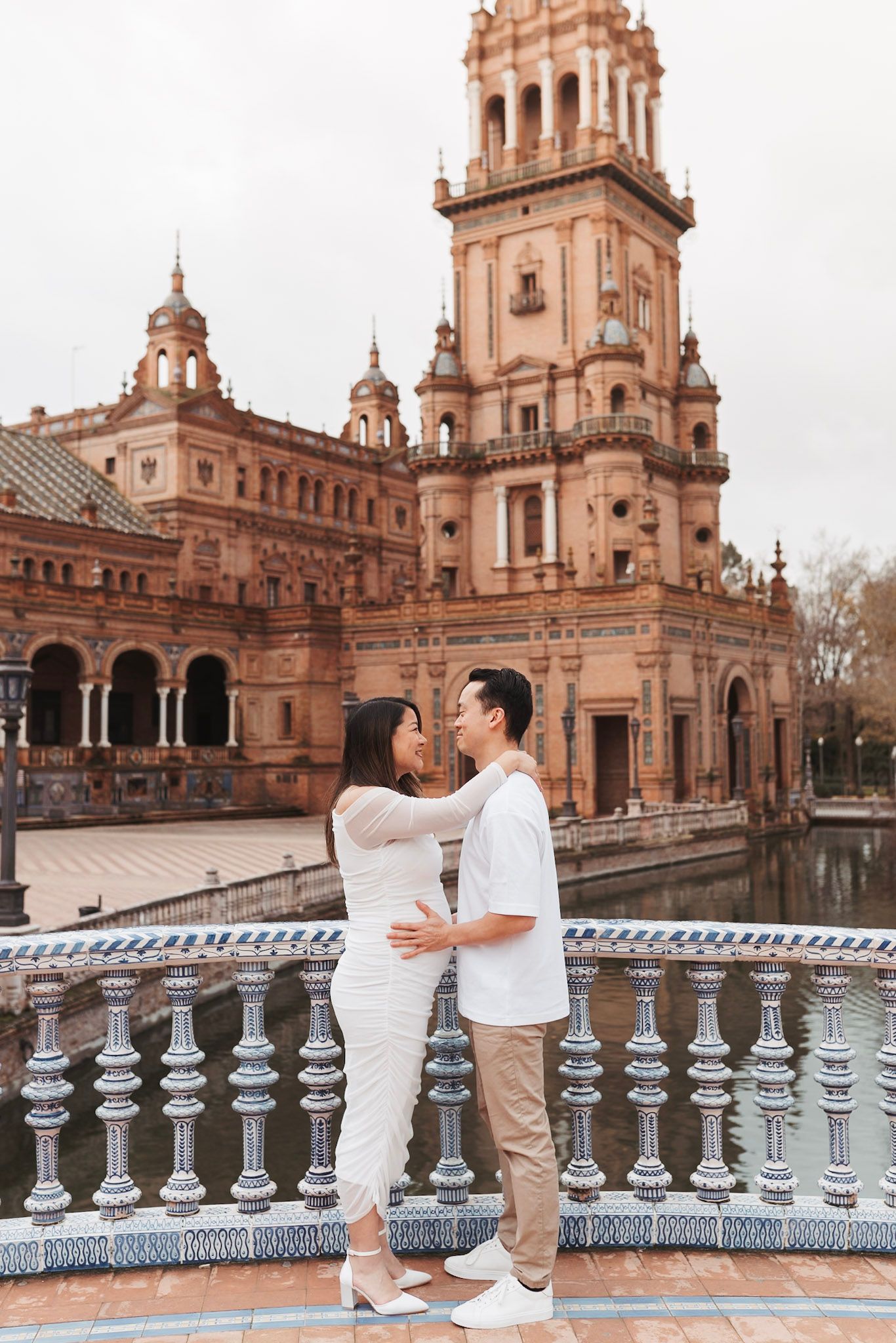 Expecting couple embraces lovingly on ornate bridge with Plaza de Espana tower in background.
