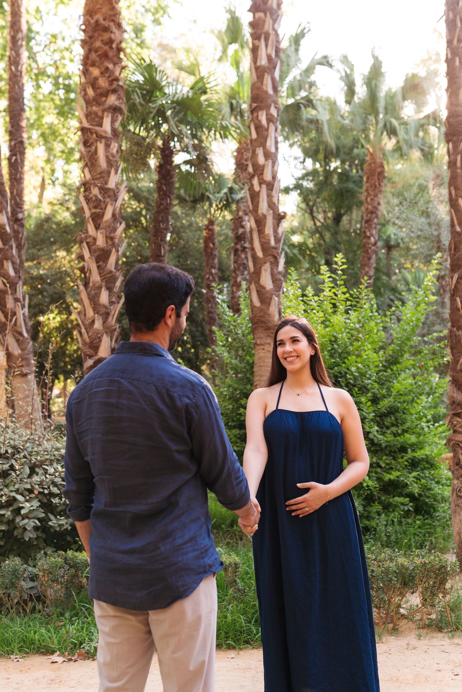 Pregnant woman in navy dress holds hands with man among palm trees outdoors.