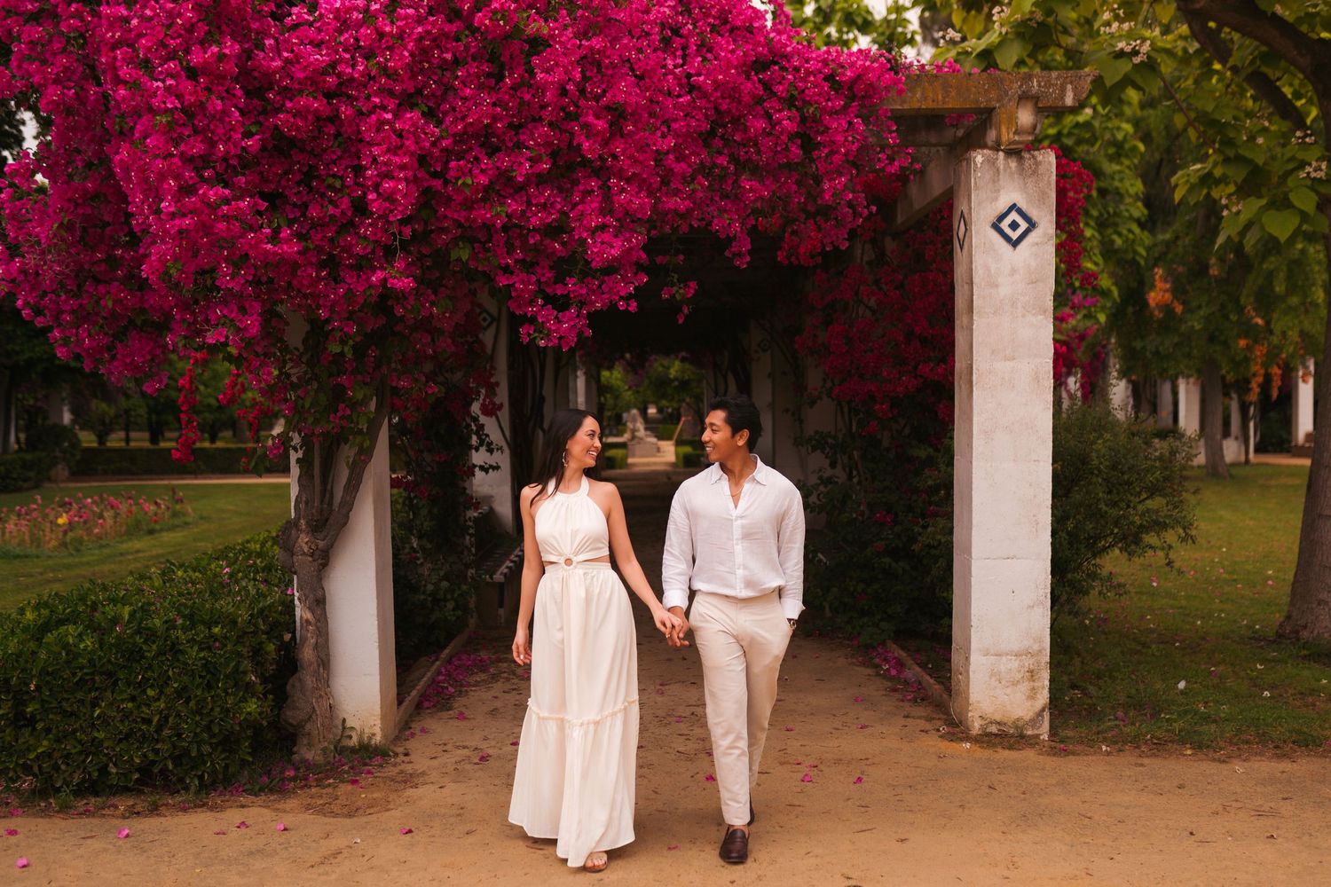 Couple walks hand-in-hand under vibrant pink bougainvillea arch on a sandy garden path.