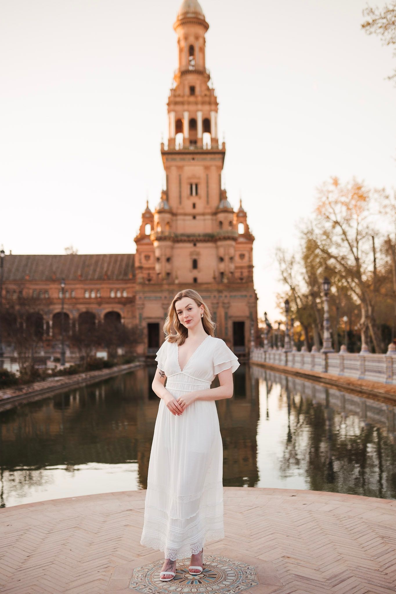 Woman in white dress stands before Plaza de España tower at sunset.