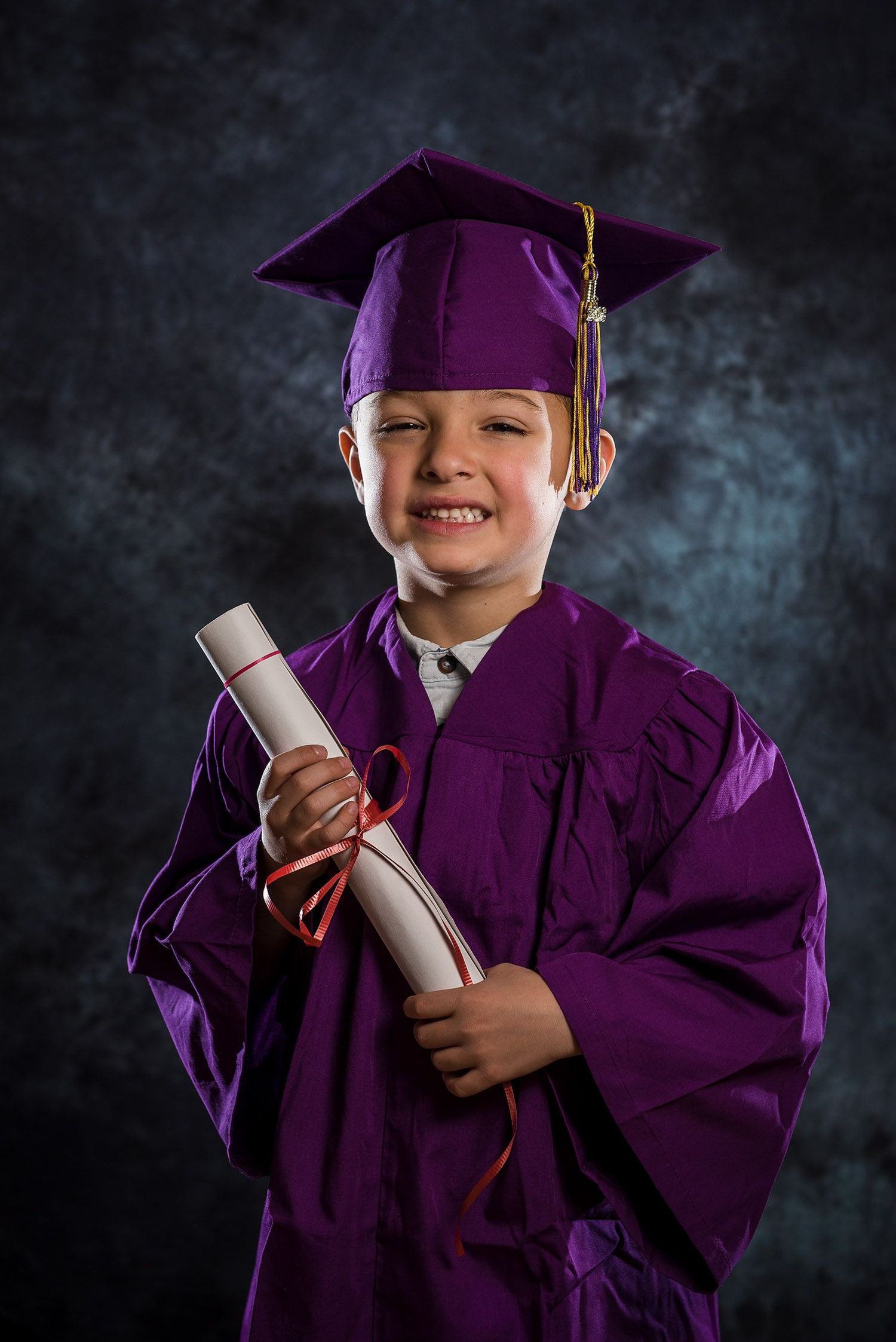 Young child in purple graduation cap and gown proudly holding a diploma scroll.