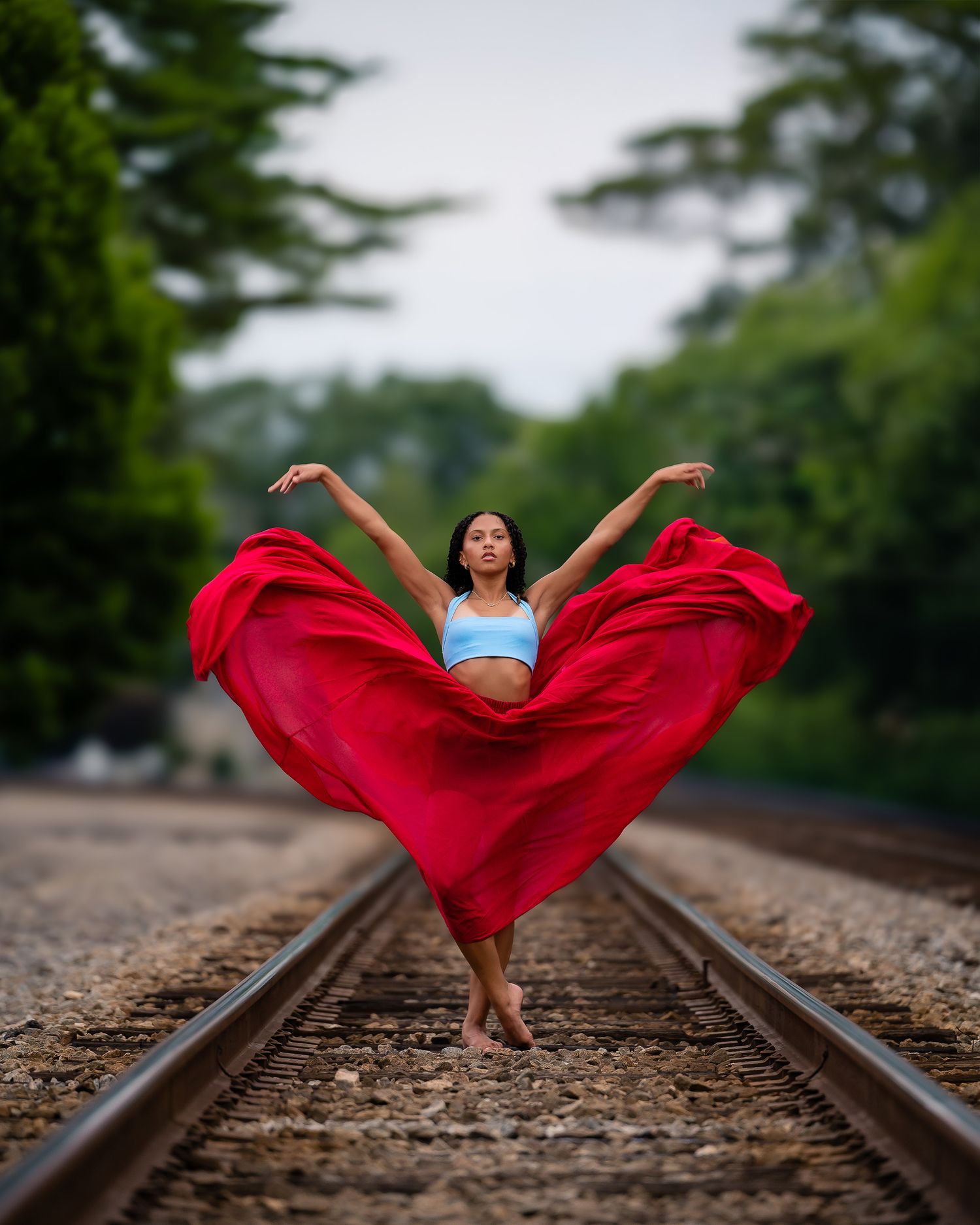 Dancer performs grand jeté on railroad tracks with flowing red skirt spread wide.