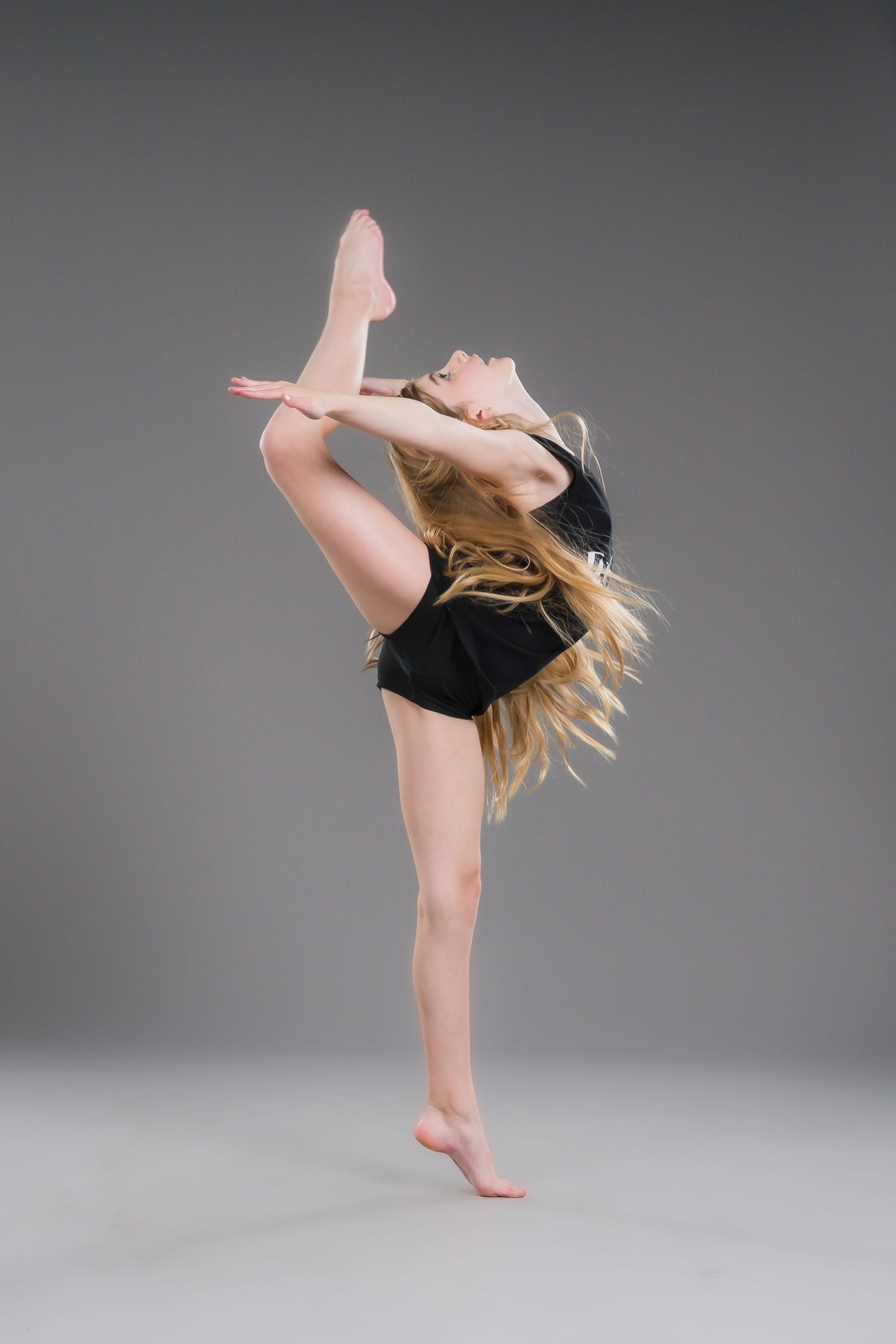 Young blonde dancer in black outfit performing high kick leap in gray studio.