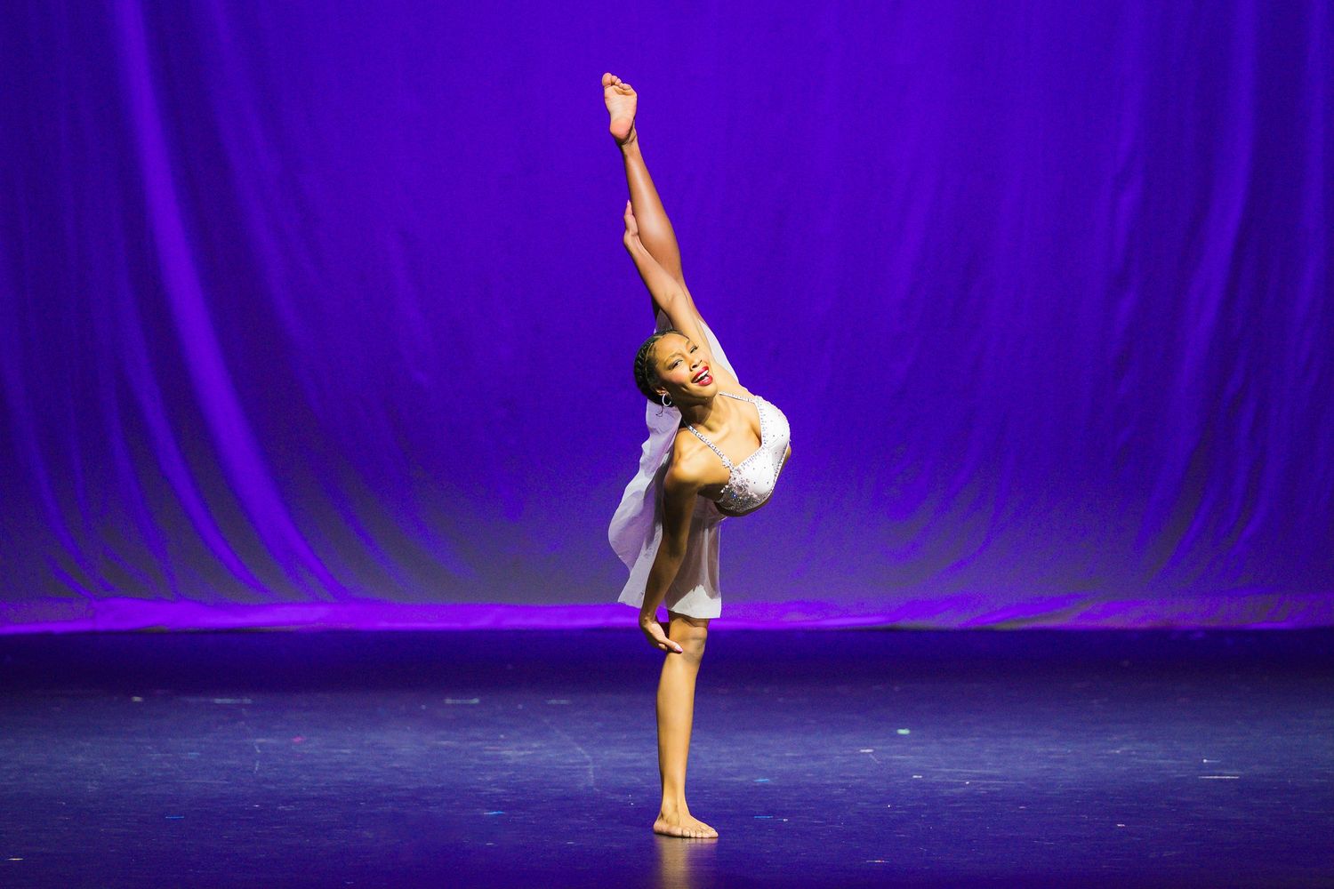 Young dancer performs an elegant arabesque on stage with a purple backdrop during a dance recital.