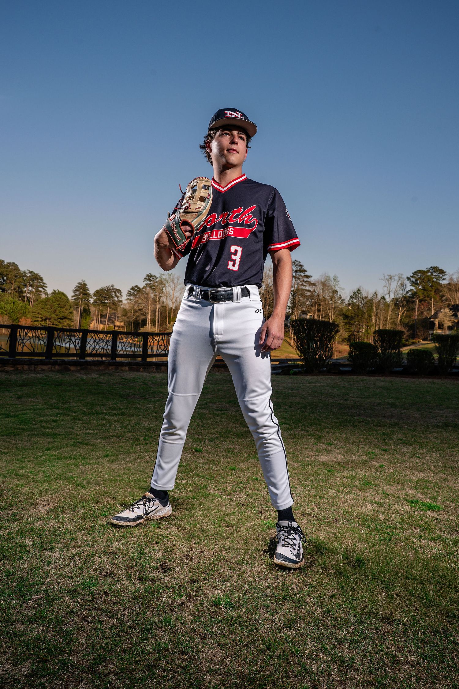 Teen baseball player in uniform number 3 holding glove on field under blue sky.