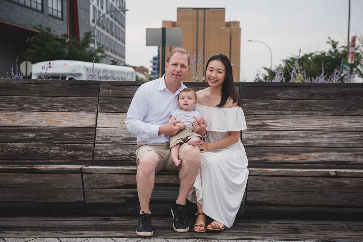 Young family with baby sitting on wooden bench outdoors in urban setting.