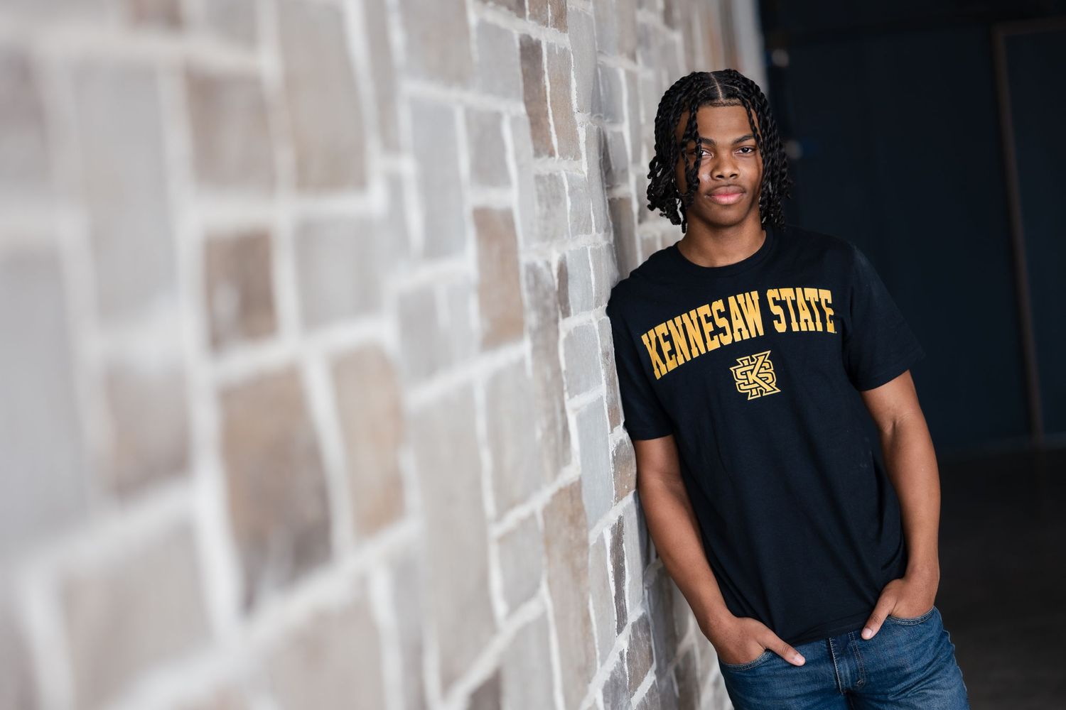 Young man in Kennesaw State t-shirt leaning against brick wall in casual portrait.