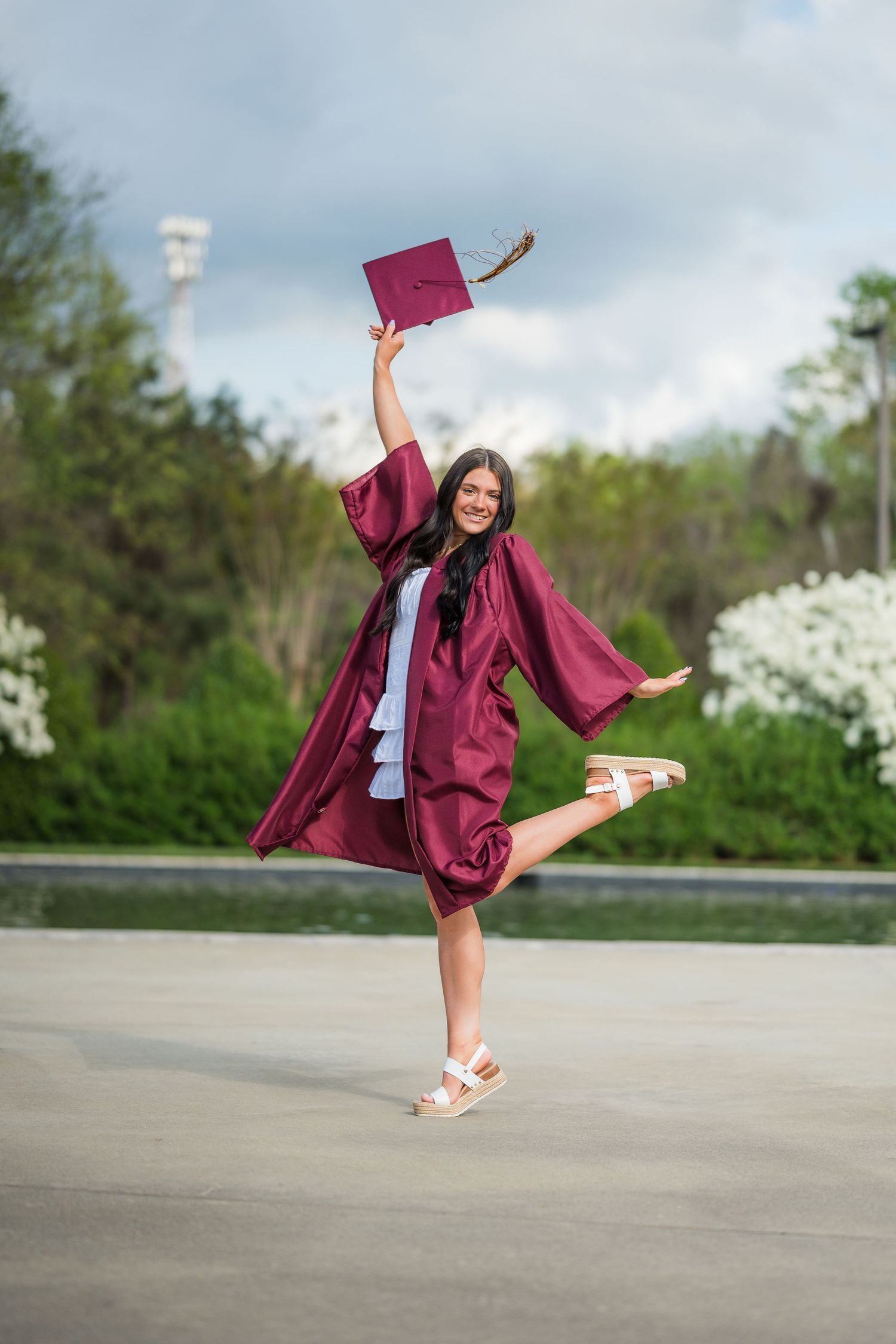 Excited graduate in maroon cap and gown celebrating outdoors by raising her cap in the air.