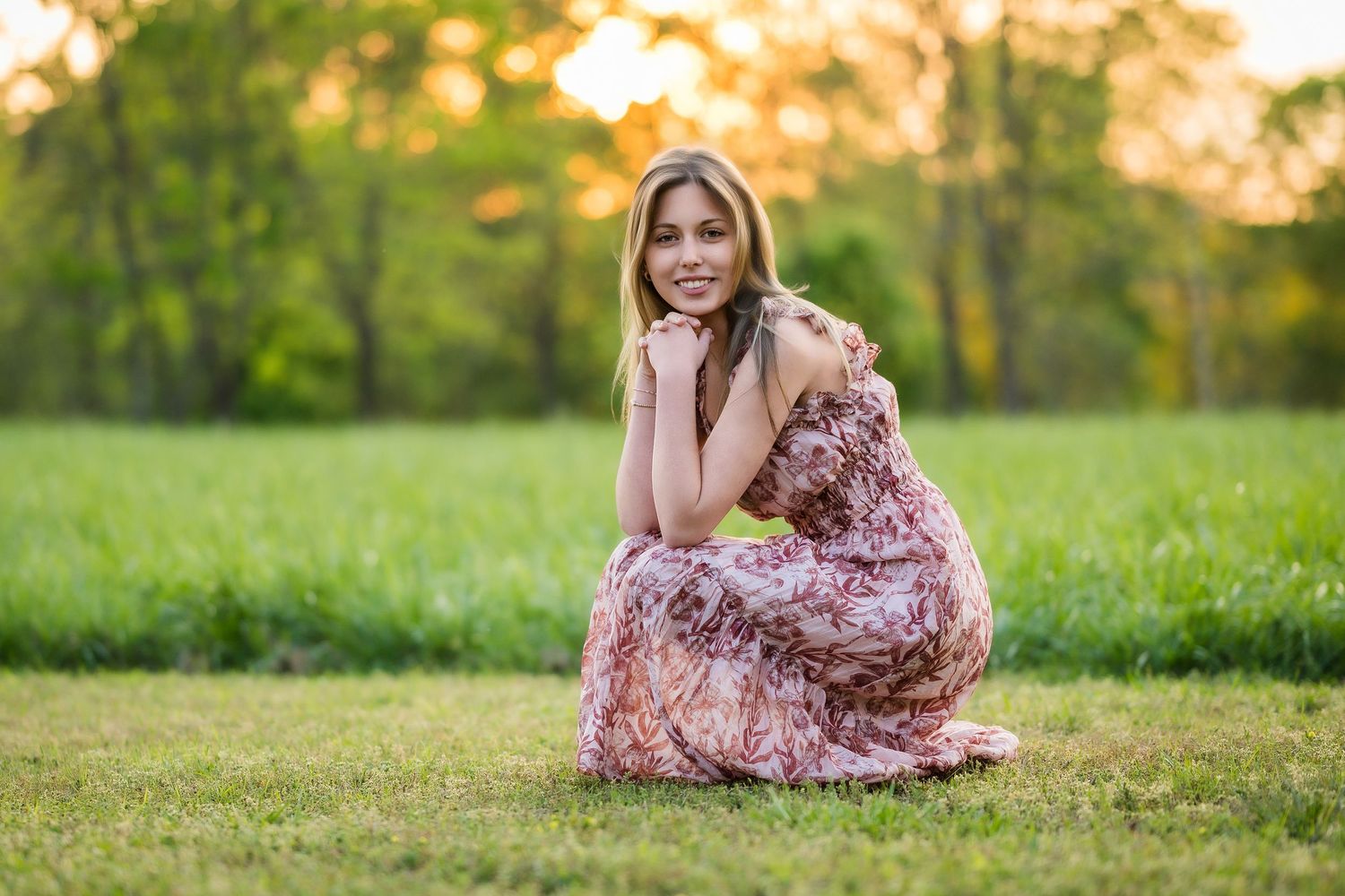 Young woman in floral maxi dress sitting gracefully on green grass field with golden sunset trees in background.