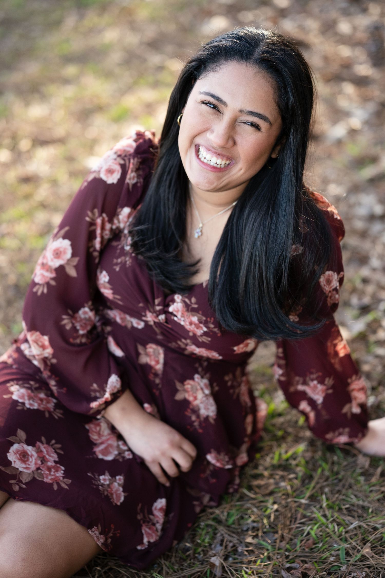Smiling Latina woman in floral burgundy dress sitting outdoors in natural setting portrait photo.