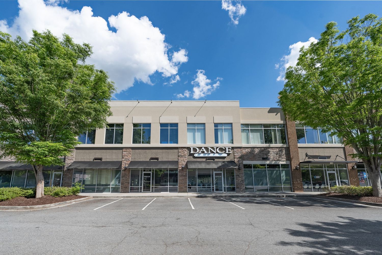 Modern two-story commercial building with Dance studio storefront, surrounded by lush green trees under a bright blue sky.