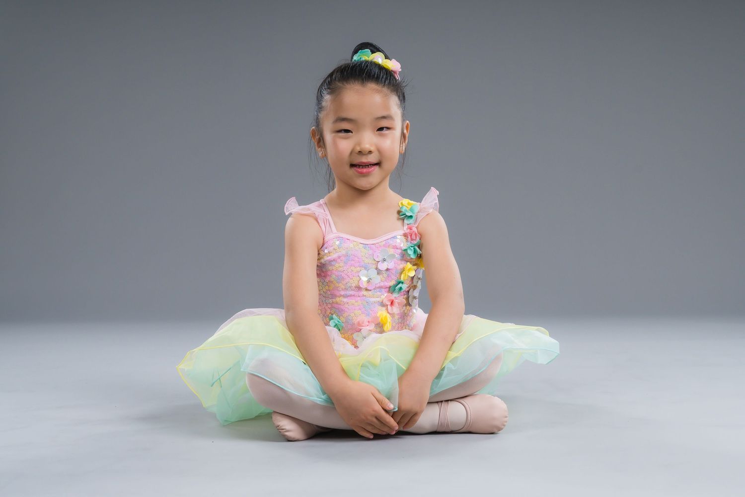 Young girl in pink ballet tutu leotard sitting cross-legged smiling on gray studio background.