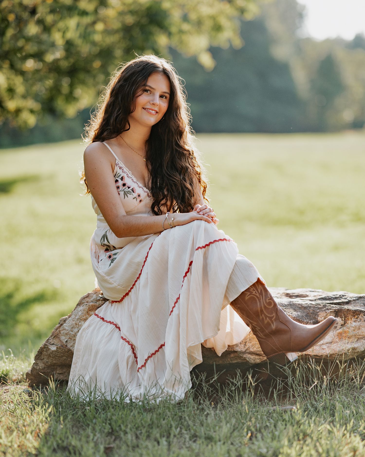Young woman in white floral dress and cowboy boots sitting on a rock in a sunny green field.