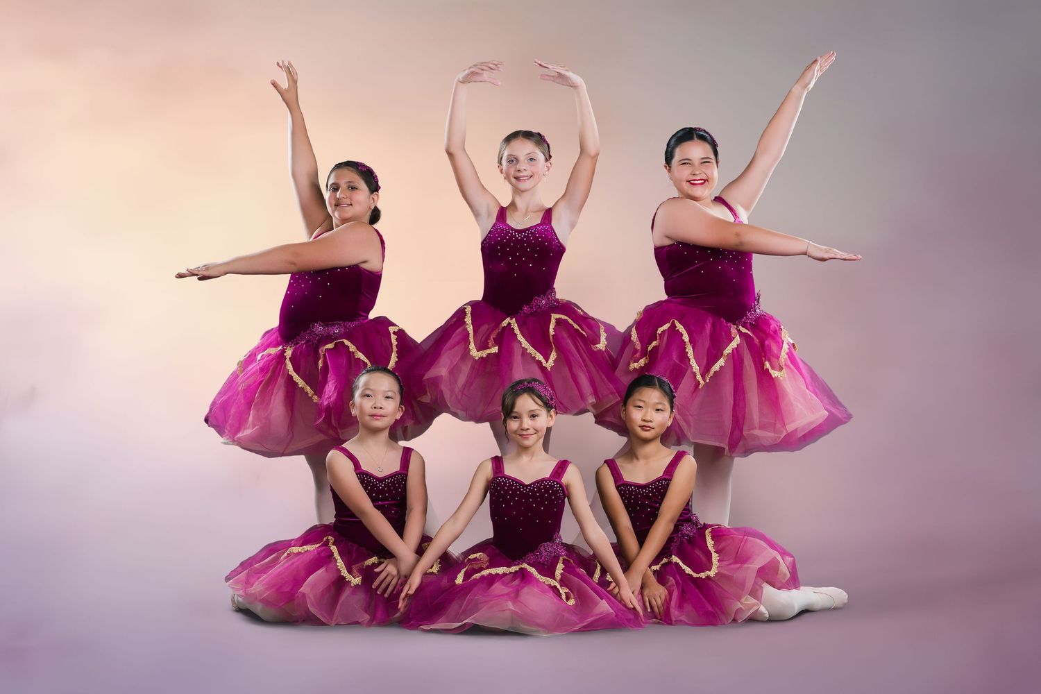 Six young ballerinas pose in pink tutus for a studio portrait against a soft pink background.