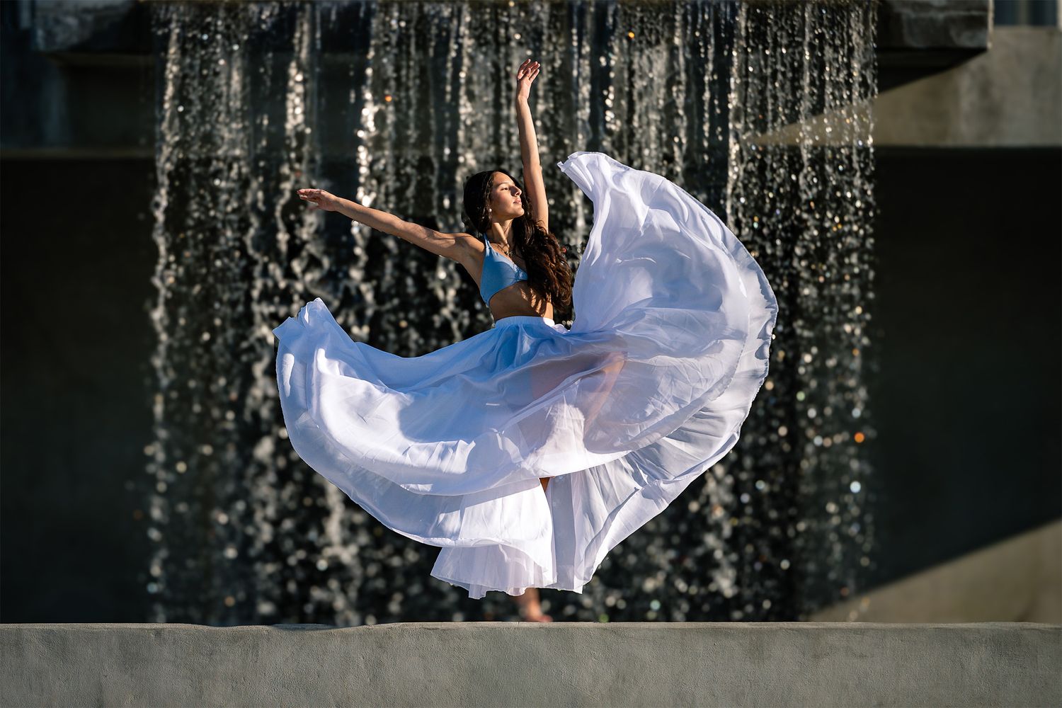 Dancer leaps gracefully in flowing white skirt near outdoor water feature.
