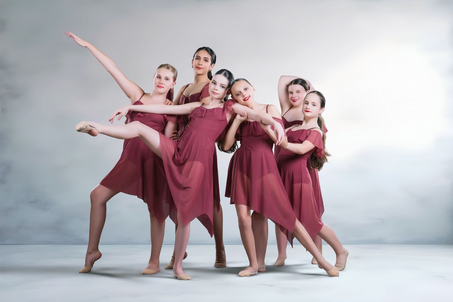 Six female dancers in matching burgundy dresses pose gracefully together in a studio setting.